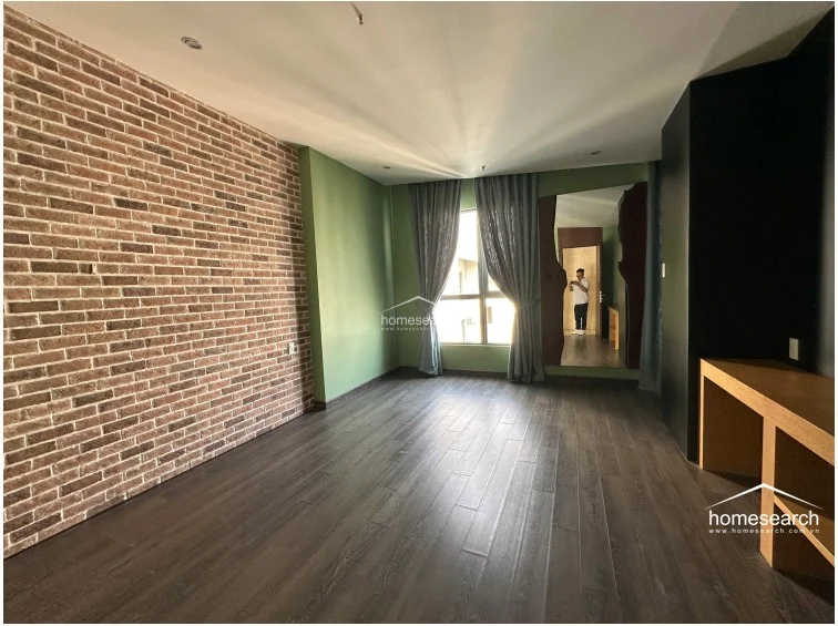 This bedroom features a striking industrial-style exposed brick feature wall contrasted by sage green paint and dark wood-look flooring. It includes a built-in wooden desk, a floor-to-ceiling mirror, and large windows with grey curtains that allow for ample natural light.