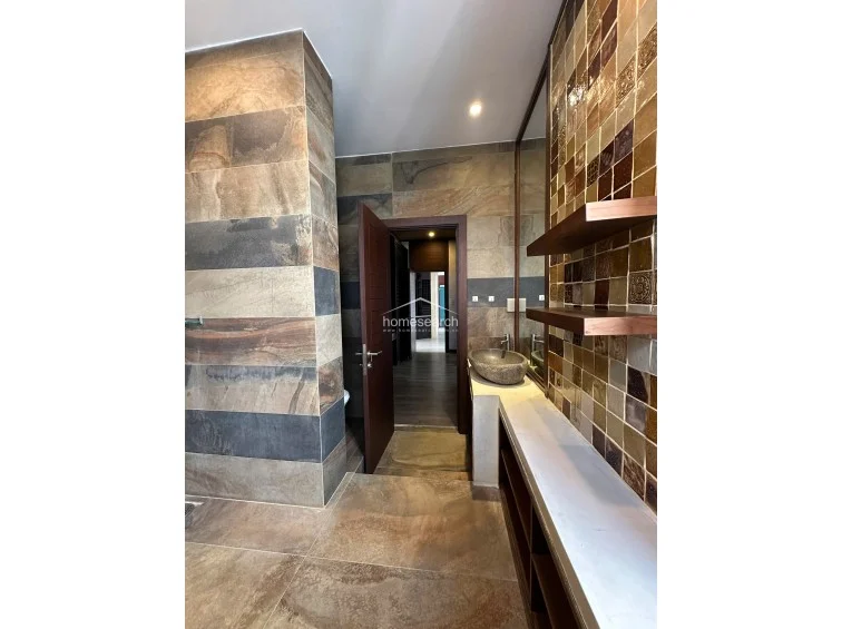 This view highlights the bathroom's high-end finishes, including the textured stone wall cladding and the warm, earthy color palette of the mosaic tiles. The space is well-lit and features a custom-built vanity area with open storage, reflecting a sophisticated and well-maintained design.