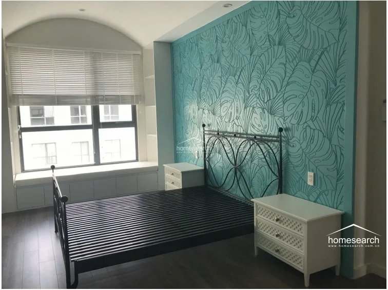 This bedroom features a distinctive arched ceiling and a vibrant teal accent wall decorated with a bold leaf-pattern mural. It includes a metal-framed bed, matching white nightstands, and a built-in window seat with storage underneath, offering a cozy and stylish retreat.
