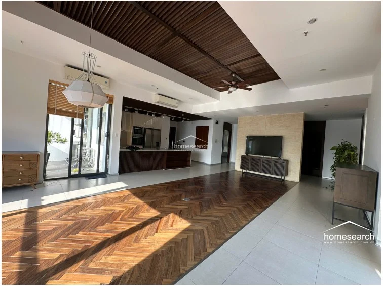 This expansive open-plan living area showcases a sophisticated mix of herringbone-patterned wood flooring and large light-colored tiles. The space features a dramatic slatted wood ceiling, a modern kitchen with a breakfast bar, and large sliding glass doors that provide seamless access to the outdoor balcony.