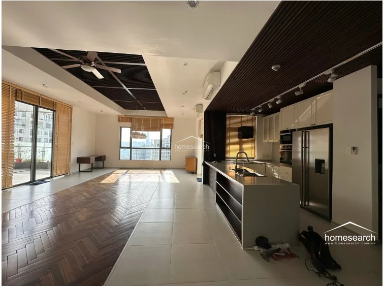 This expansive open-concept living and kitchen area showcases a striking contrast between herringbone-patterned wood flooring and neutral tile zones. The space is defined by high ceilings with dark wood slat detailing, integrated lighting, and large windows that offer significant natural light and urban views.