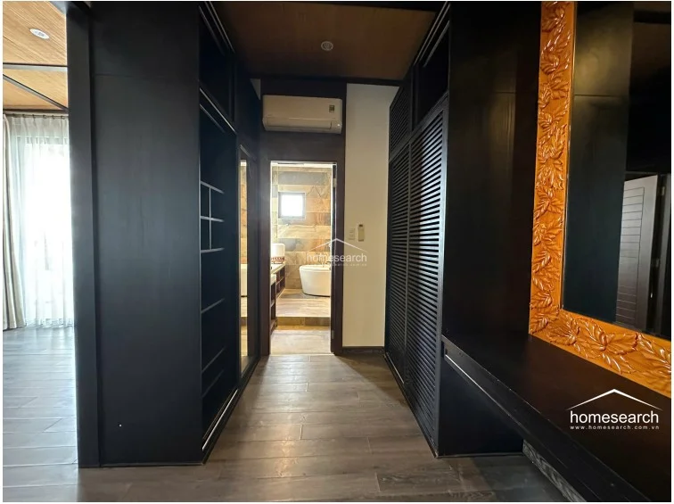 This walk-in closet area features extensive dark wood cabinetry with louvered doors, providing ample storage and a sleek, modern aesthetic. It serves as a transitional space leading into a bathroom, which is visible in the background with stone-tiled walls and a soaking tub.