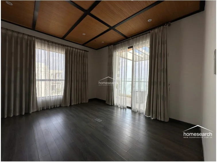 This bedroom mirrors the design of the previous one, showcasing dark wood flooring and a textured, grid-patterned ceiling. The room is framed by large windows with layered curtains that open onto a private balcony, maintaining a consistent, well-maintained aesthetic.