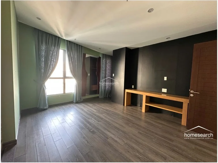 This bedroom features a bold color scheme with sage green walls contrasted against a striking black accent wall. It includes a custom built-in wooden console desk, dark wood flooring, and a large window dressed in floor-length curtains that allow for plenty of natural light.