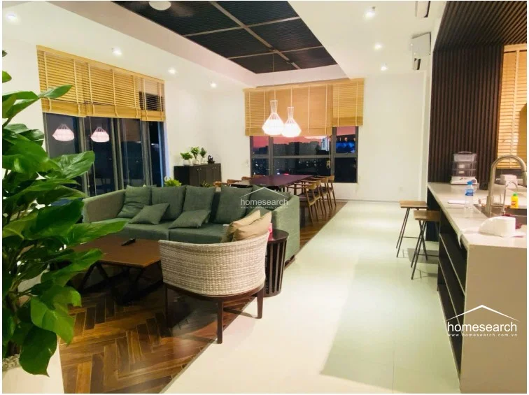 This open-concept living and dining area boasts a sophisticated blend of herringbone wood flooring and light-colored tiles, creating a distinct visual separation. The room is flooded with natural light from floor-to-ceiling windows, complemented by modern pendant lighting and a sleek, dark-toned kitchen island with bar seating.