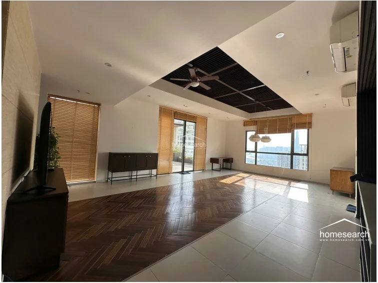 This expansive living area features a sophisticated mix of herringbone-patterned wood flooring and light-toned tile, creating a distinct visual separation between zones. The room is bathed in natural light from large windows and a glass balcony door, both dressed with bamboo blinds, while a dark-toned recessed ceiling with a central fan adds architectural interest.