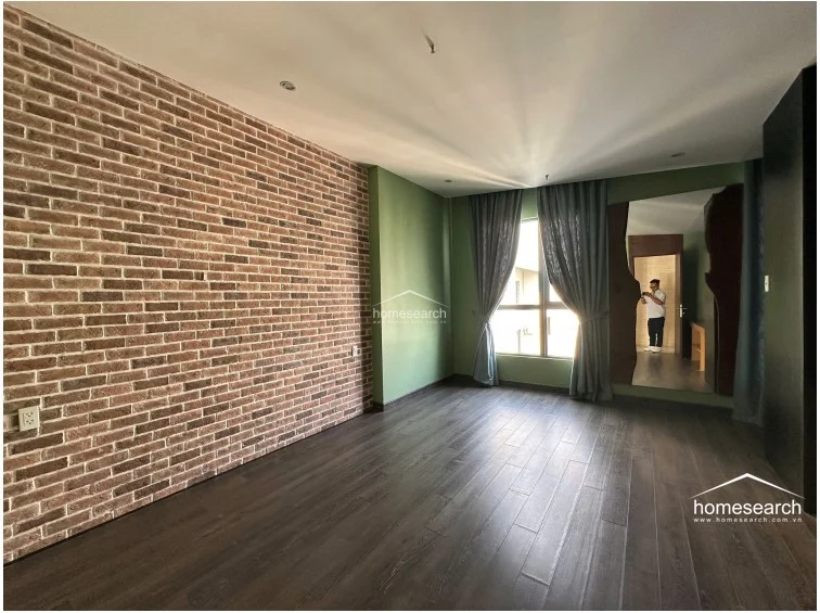 This spacious bedroom features a striking exposed brick feature wall paired with olive green painted walls and dark wood-look flooring. It includes a large window with floor-to-ceiling curtains and a full-length mirror, creating a modern, industrial-inspired aesthetic that appears to be in excellent condition.