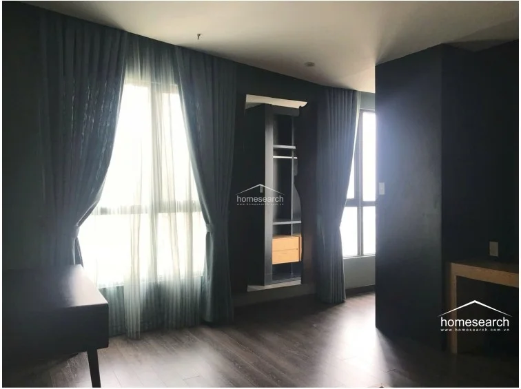 This minimalist bedroom is designed with a moody, dark color palette and rich wood flooring, creating a serene and private retreat. It includes a built-in open-concept wardrobe system and large windows dressed in floor-length drapes that allow for ample natural light.