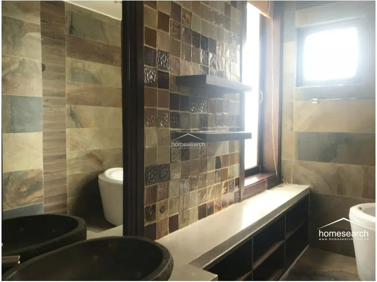 This bathroom features a unique design with decorative mosaic wall tiles and earthy, multi-tonal stone-look floor tiles. It is equipped with a modern vessel sink, a long vanity with open storage shelving, and a window that provides natural light while maintaining privacy.