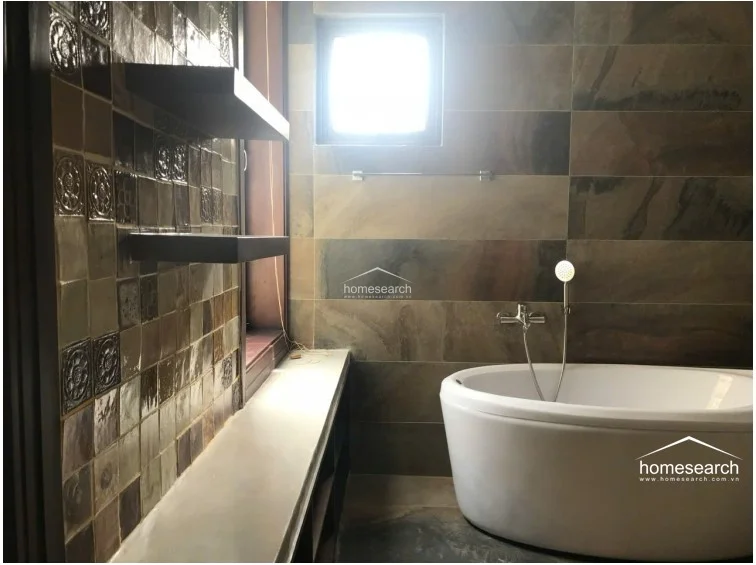 This bathroom features a sophisticated design with large-format slate-look wall tiles and a striking accent wall of textured, dark-toned decorative tiles. A modern white freestanding soaking tub serves as the focal point, complemented by a built-in stone ledge and floating shelves that provide functional storage. The space is well-maintained and includes a small window that allows for natural ventilation and soft light.