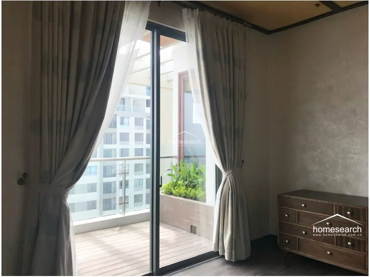 This bedroom offers a seamless transition to a private outdoor balcony through full-height sliding glass doors, which are dressed in elegant floor-to-ceiling curtains. The room features a warm, textured wall finish and dark wood-toned flooring, paired with a stylish mid-century modern wooden dresser. The balcony includes wood-look decking and a built-in planter box, creating a serene, well-integrated indoor-outdoor living experience.