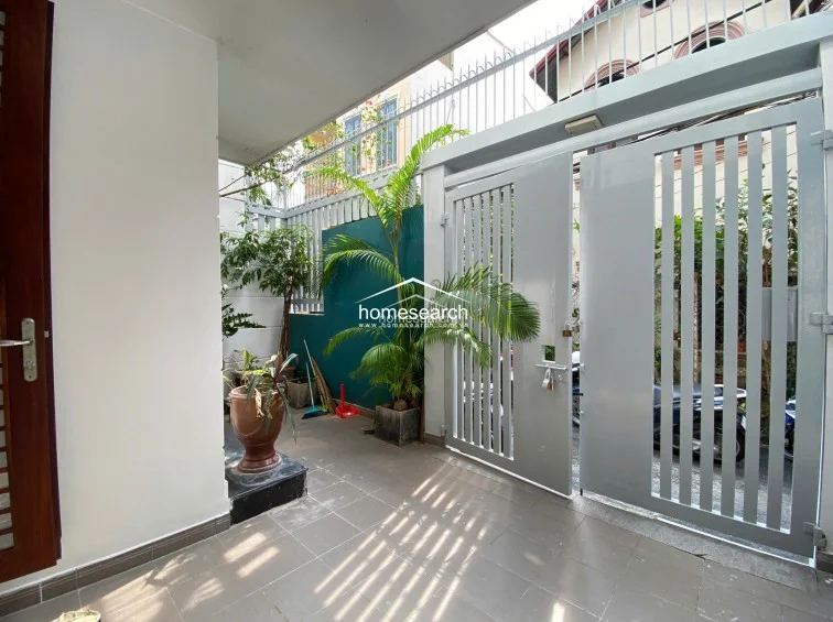 This view from the terrace looks toward the street, highlighting the secure metal gate and a vibrant green accent wall. The area is well-maintained and serves as a functional outdoor foyer, offering a small, shaded space for potted plants and storage.
