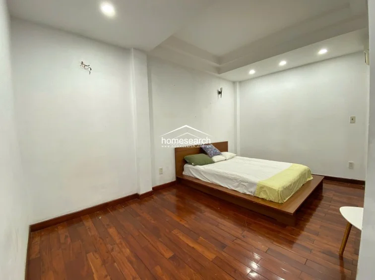This bedroom features warm-toned hardwood flooring and a minimalist platform bed, creating a cozy and clean aesthetic. The space is illuminated by recessed ceiling lighting and features a simple, neutral color scheme that maximizes the sense of openness.