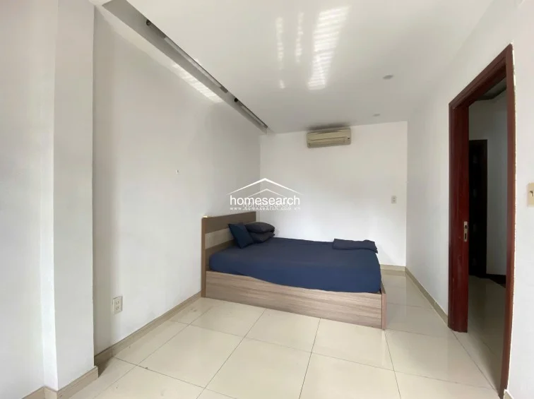 This bedroom is finished with light-colored floor tiles and includes a wall-mounted air conditioning unit for climate control. The room is accessed via a dark wood door frame, providing a sharp contrast against the plain white walls.