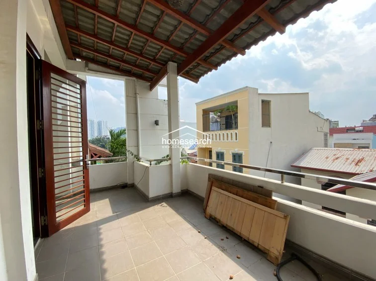 This spacious balcony features a covered roof with exposed wooden beams and provides an open-air view of the surrounding neighborhood. It is finished with neutral floor tiles and a stainless steel railing, offering a functional outdoor space for relaxation or storage.