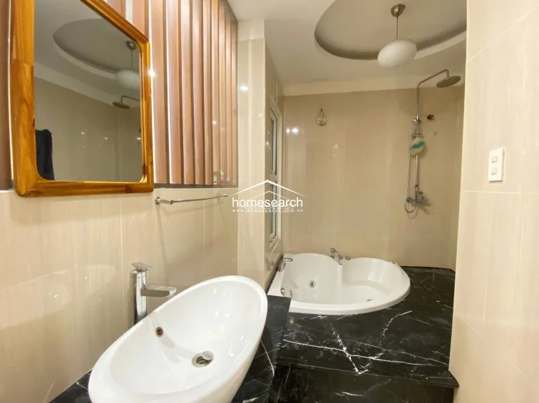 This bathroom highlights a luxurious corner soaking tub paired with a separate rainfall showerhead, creating a spa-like atmosphere. The space is accented by a warm wooden slat feature wall, a stylish vessel sink on a black marble vanity, and a recessed ceiling light for a sophisticated finish.