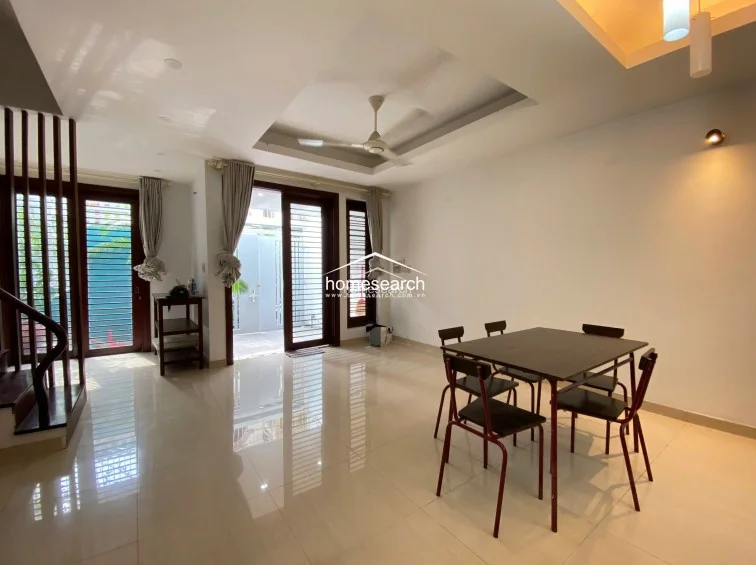 This open-plan living and dining area features polished light-colored tile flooring and a clean, white color scheme that enhances the sense of space. A central wooden staircase with decorative balusters serves as a focal point, while the recessed ceiling with a fan provides a modern architectural touch.