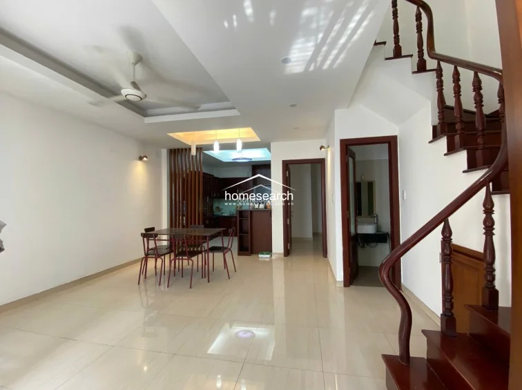 This shot reveals the full layout of the ground floor, showing the dining area transitioning into a kitchen space partially screened by wooden slats. The inclusion of a ground-floor bathroom and a prominent wooden staircase indicates a well-organized, functional floor plan with consistent flooring throughout.