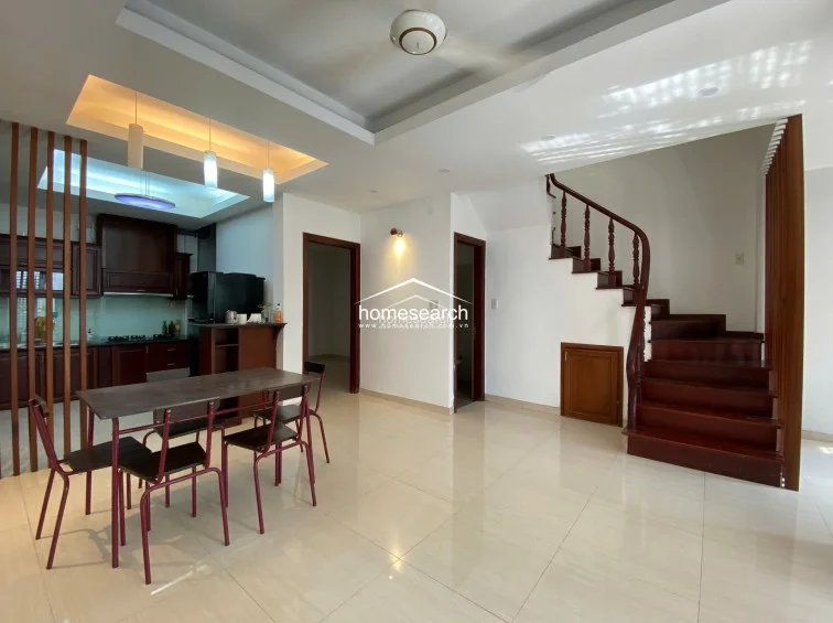 This open-concept living and dining area features a modern kitchen with dark wood cabinetry and a breakfast bar. The space is anchored by a polished tile floor and a distinctive wooden staircase with a classic balustrade, connecting the ground floor to the upper levels.