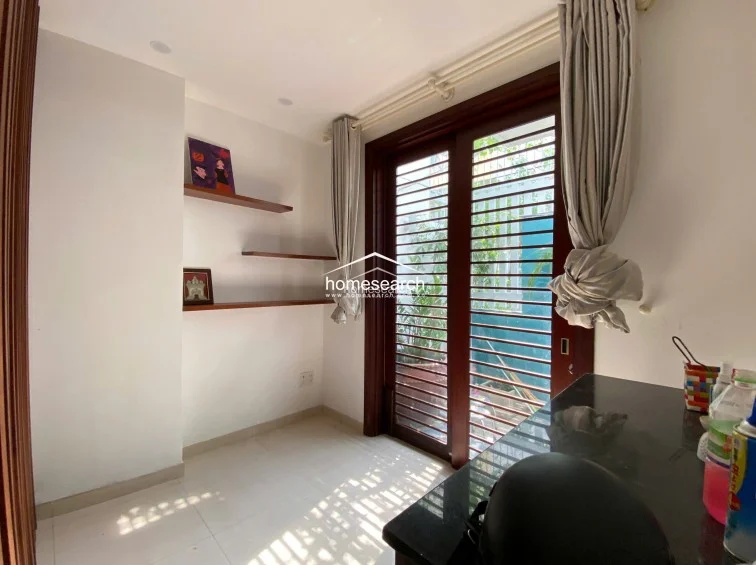 This compact, multi-purpose room functions as a study or workspace, featuring built-in wooden shelving and a sleek black desk surface. Natural light streams in through a large glass-paned door that provides access to an outdoor area, while neutral walls and tile flooring maintain a clean, minimalist aesthetic.