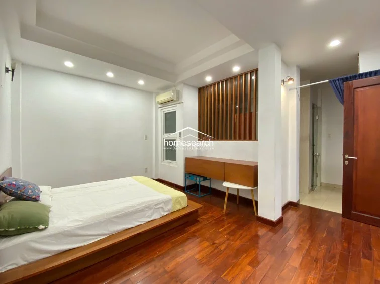 This bedroom features warm, rich hardwood flooring and a low-profile platform bed, creating a cozy and contemporary atmosphere. The room includes a wall-mounted AC unit, a built-in wooden console, and a decorative slatted room divider that adds texture and visual interest to the space.