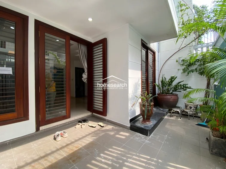 This image captures a private, paved outdoor terrace or entryway featuring large ceramic planters and lush greenery. The space is enclosed by a high wall and a metal gate, providing a secure and serene transition area that connects directly to the interior through dark wood-framed glass doors.