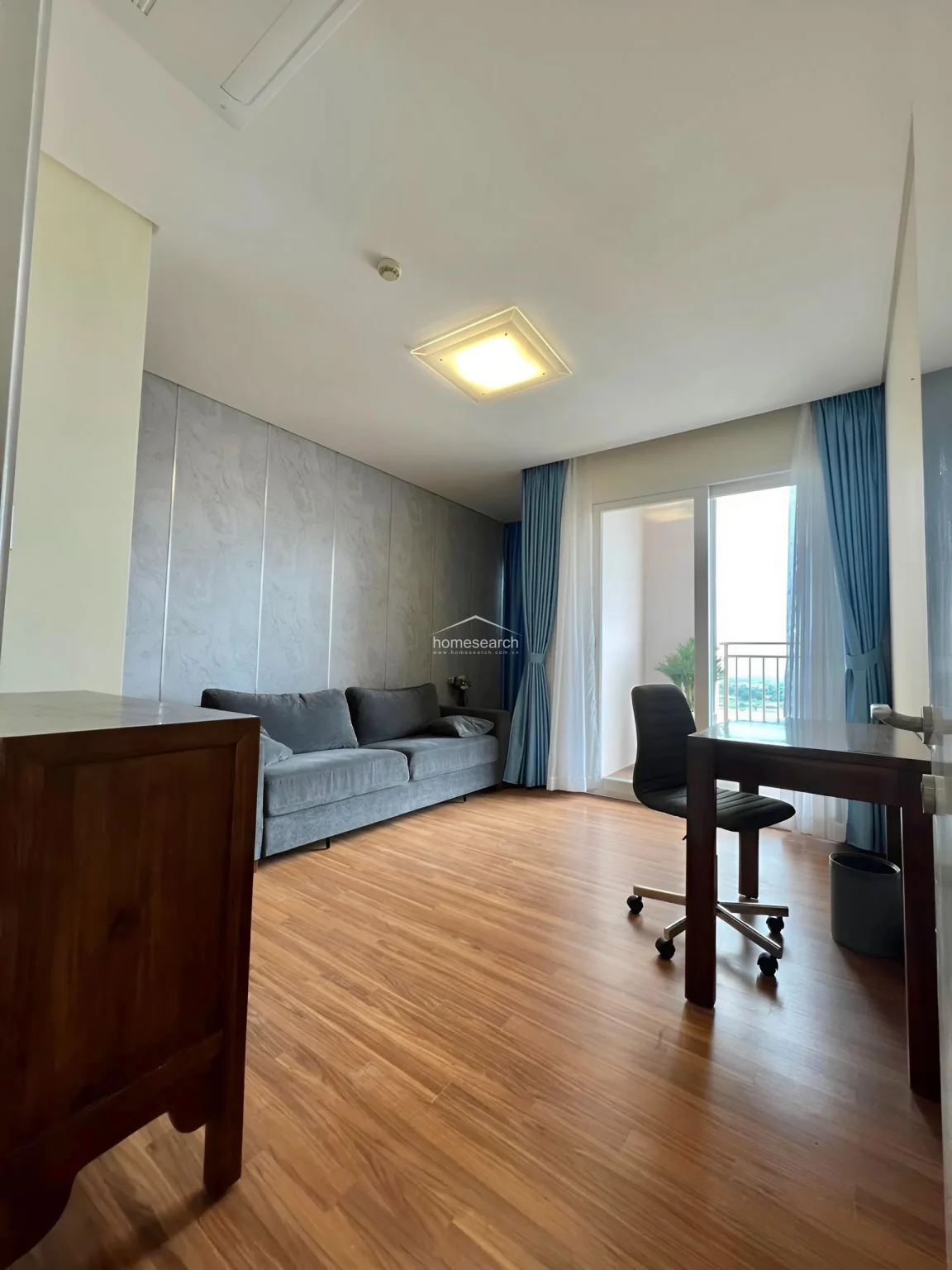 This perspective of the same room highlights the natural light entering through a balcony door, which is framed by elegant blue and white curtains. The layout provides a functional workspace and relaxation area, maintaining a clean, modern aesthetic with neutral wall tones.
