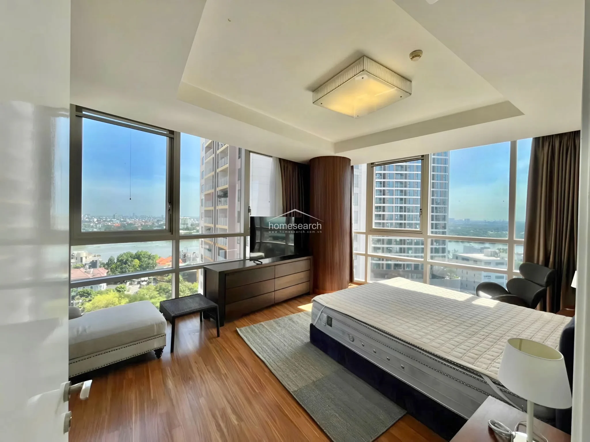 This spacious bedroom features floor-to-ceiling windows on two walls, offering abundant natural light and panoramic river views. The room is finished with warm wood-look flooring, a modern square ceiling light, and a sleek, dark wood media console, creating a sophisticated and airy atmosphere.