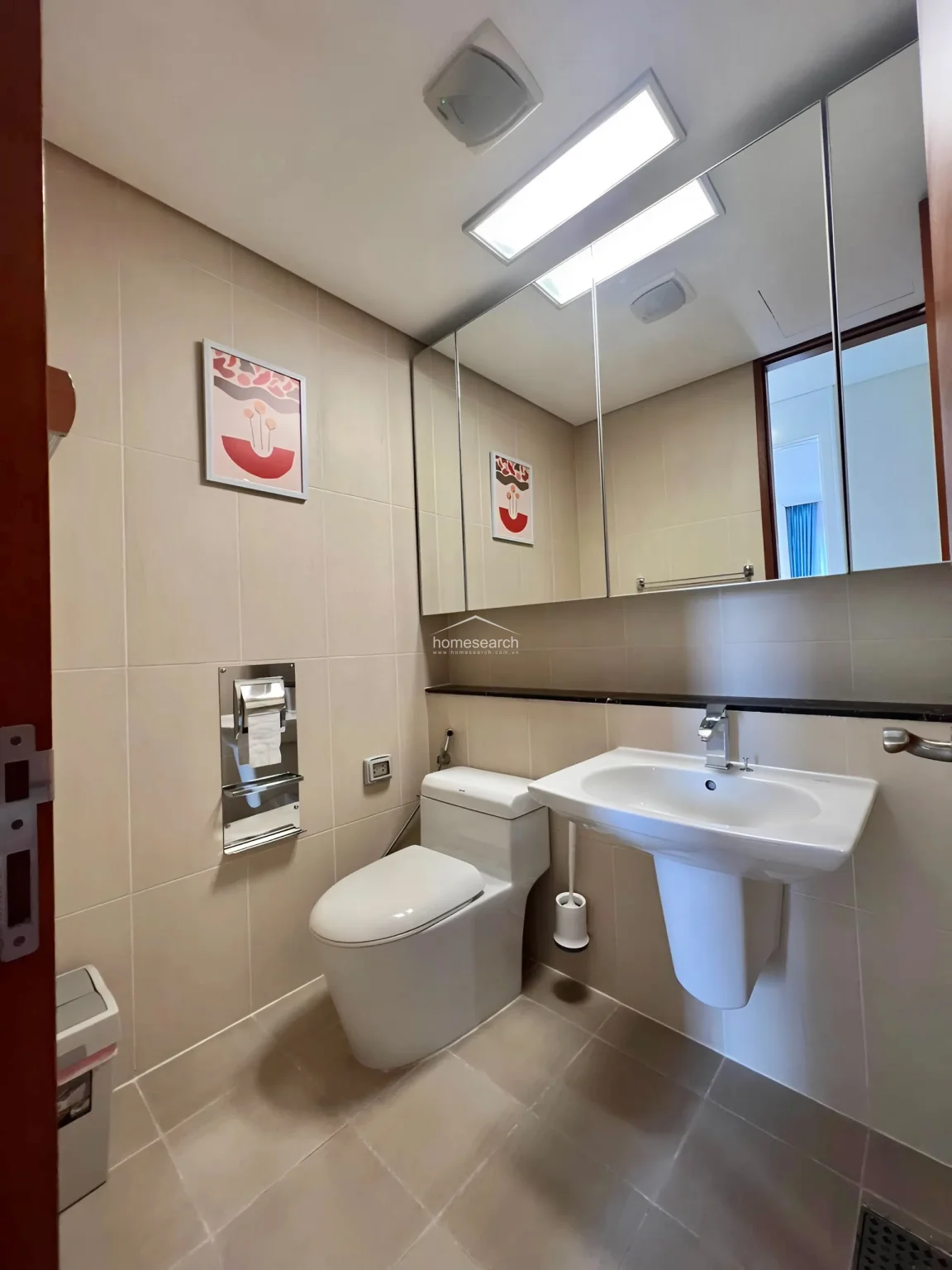 This compact powder room or secondary bathroom is finished in neutral beige tiles, offering a clean and minimalist look. It includes a wall-mounted pedestal sink, a modern toilet, and a large, integrated mirror cabinet that provides essential storage while enhancing the sense of space.