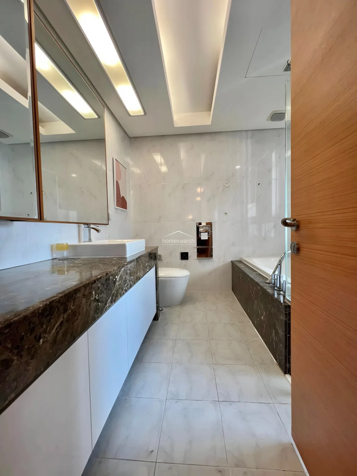 This elegant bathroom showcases a premium design with marble-topped vanity, white marble-look wall tiles, and recessed ceiling lighting. The space is in excellent condition, featuring a seamless flow between the vanity, toilet, and bathtub area, all accented by warm wood-toned doors and trim.