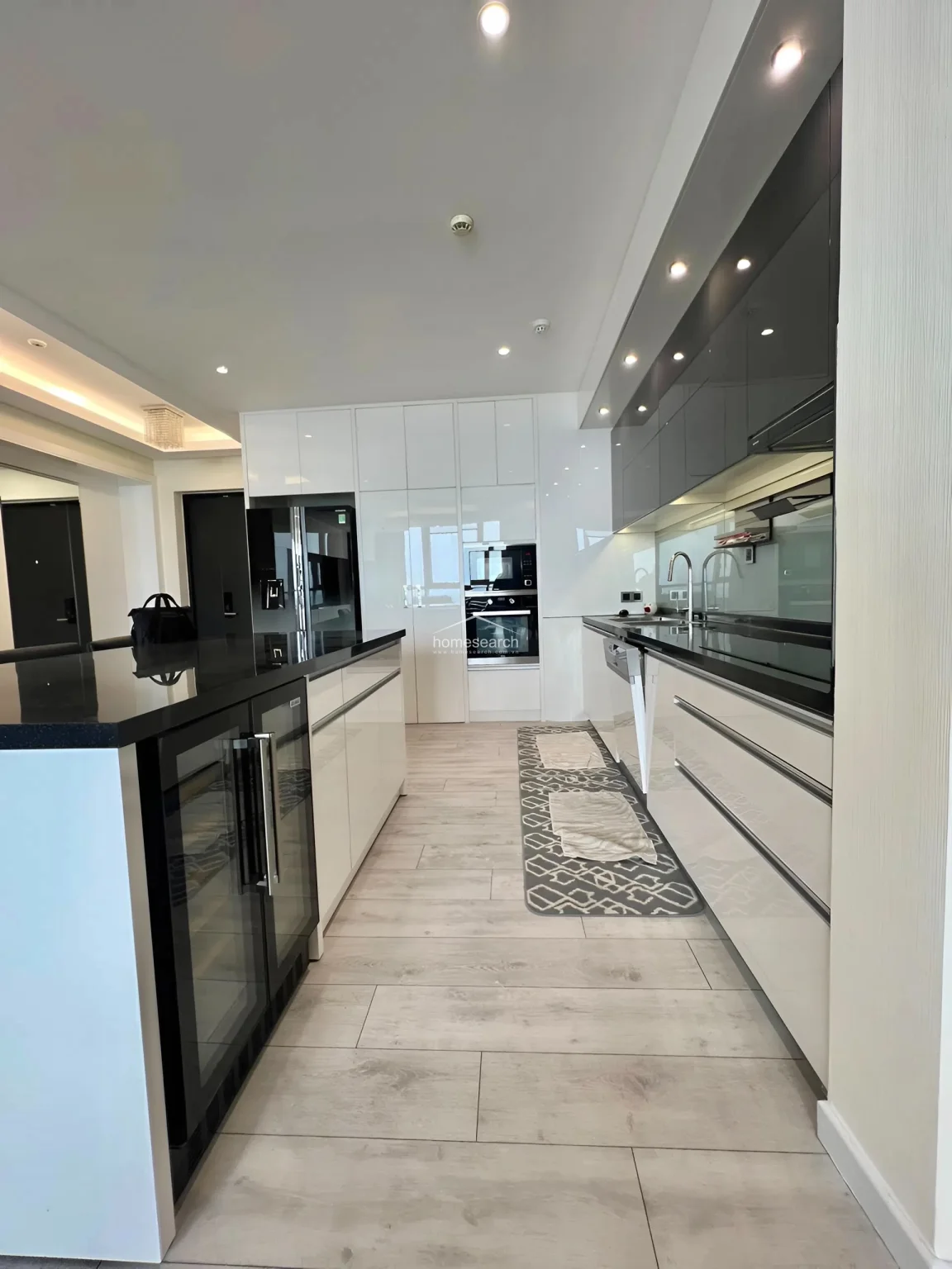 This sleek, modern kitchen features high-gloss white cabinetry, a contrasting black countertop, and an integrated wine cooler in the island. The space is well-equipped with built-in appliances, including an oven and microwave, and is finished with light-colored tile flooring for a polished, low-maintenance look.