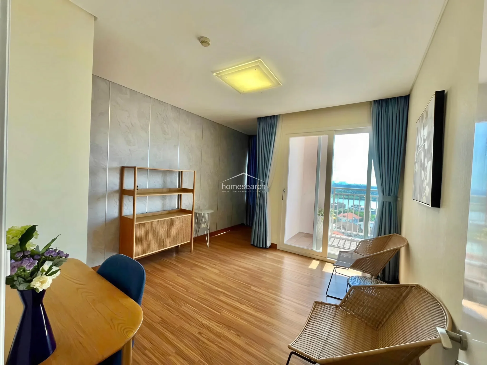 This versatile living or multipurpose room is characterized by a modern grey accent wall and warm wood flooring. The space is furnished with a light wood shelving unit and two rattan-style chairs, with large sliding glass doors that open onto a private balcony, ensuring a seamless indoor-outdoor flow.