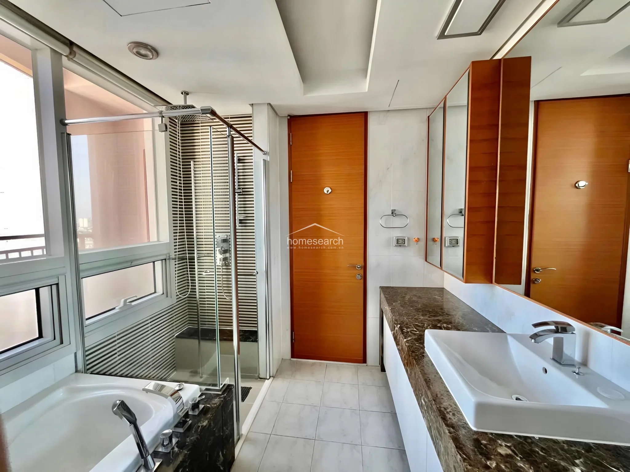 This master-style bathroom features a luxurious layout with a deep soaking tub and a separate glass-enclosed shower area. The space is finished with high-end dark marble countertops, white tile flooring, and warm wood-toned cabinetry, creating a sophisticated and well-maintained aesthetic.