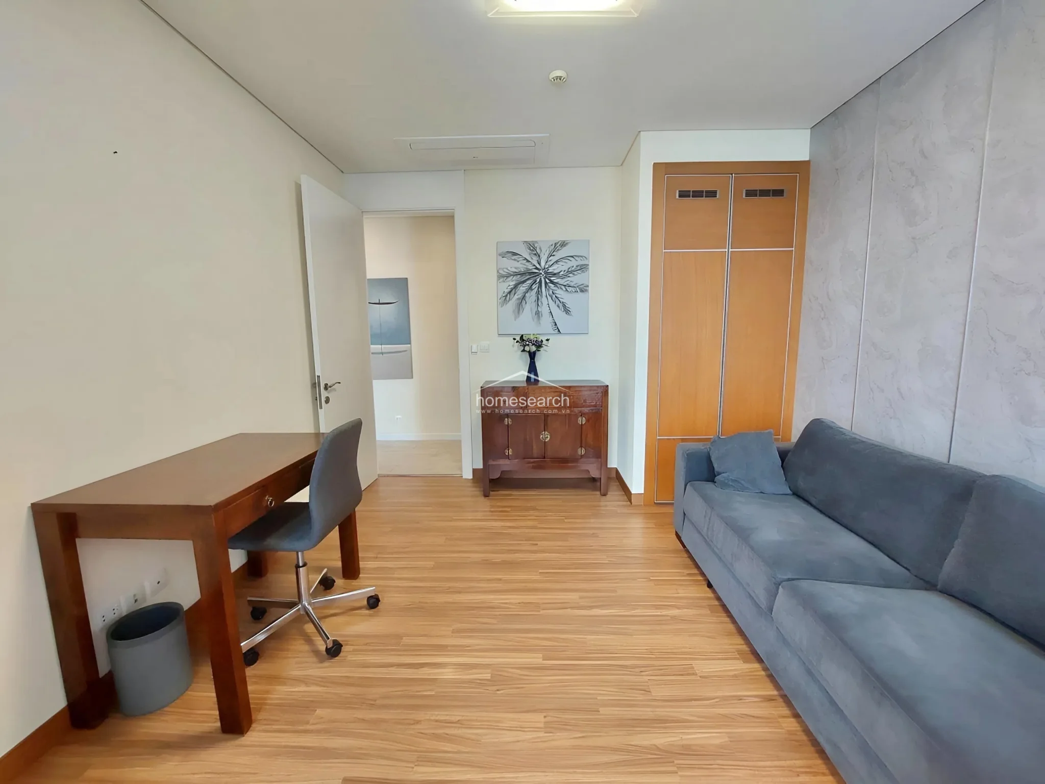 This multi-purpose room serves as a home office and lounge area, featuring warm wood-look flooring and a textured grey accent wall. It includes a built-in wooden wardrobe, a desk with a chair, and a comfortable grey sofa, with a doorway leading to an adjacent space.