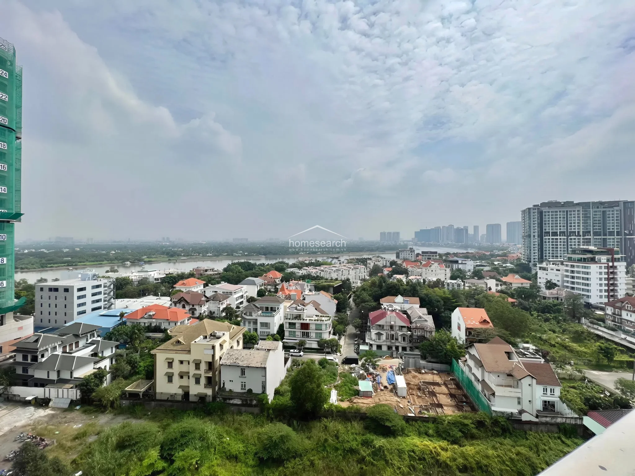 This high-elevation exterior shot showcases a sprawling urban and suburban landscape with a prominent river view. The scene highlights a mix of low-rise residential villas and taller apartment complexes, indicating a well-developed, scenic neighborhood with ongoing local construction.