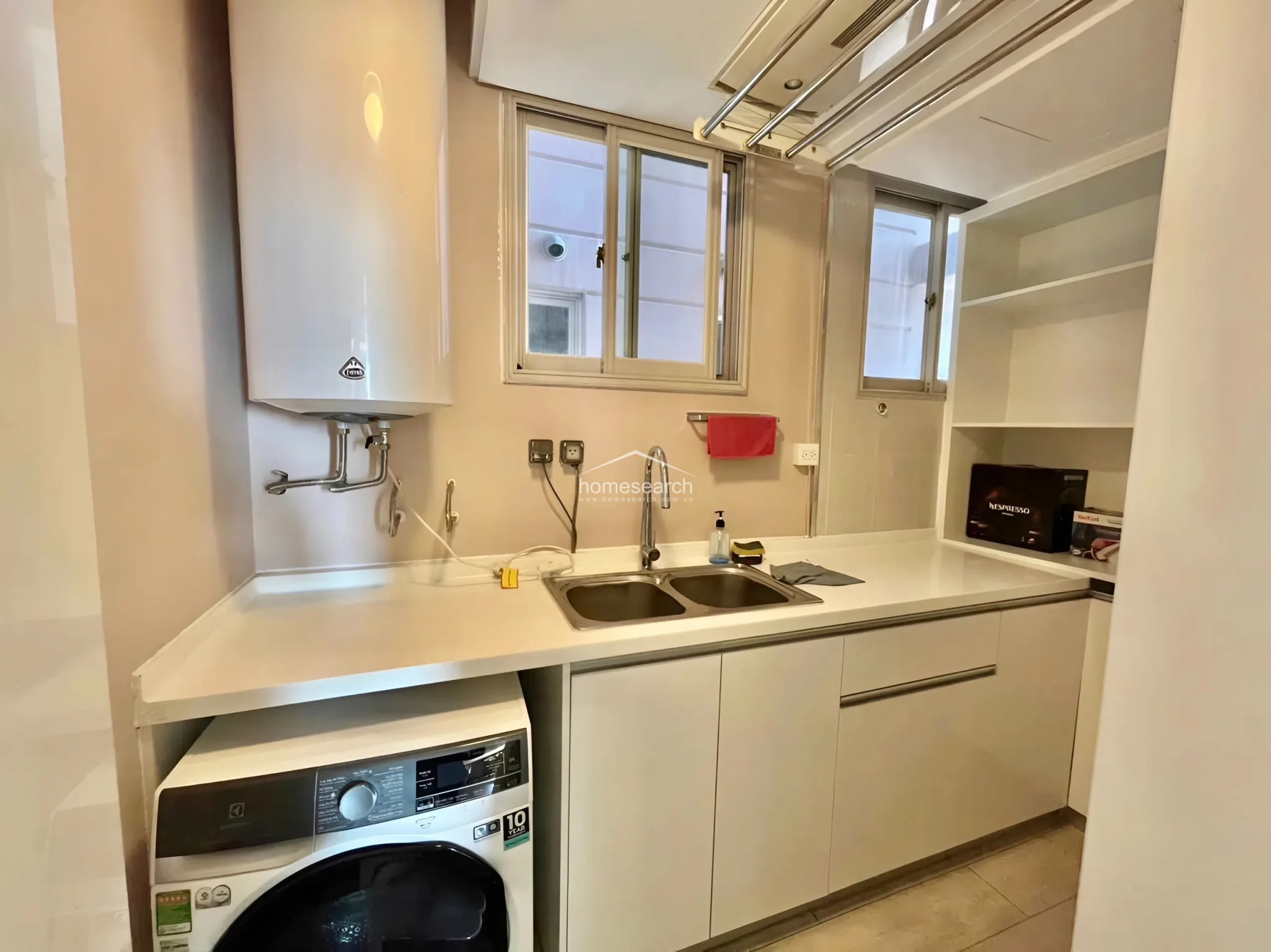 This utility or laundry area is highly functional, featuring a white washing machine, a double-basin stainless steel sink, and an overhead water heater. It includes built-in open shelving for storage and a ceiling-mounted drying rack, making it a practical and efficient space.