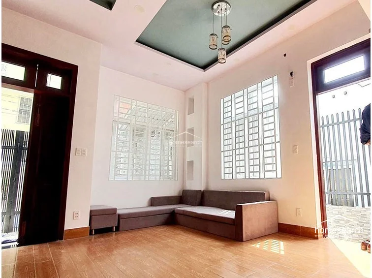 This living area features a minimalist layout with a low-profile sectional sofa and warm wood-look tile flooring. The space is brightened by large windows with security grilles and a decorative recessed ceiling with a modern pendant light fixture.