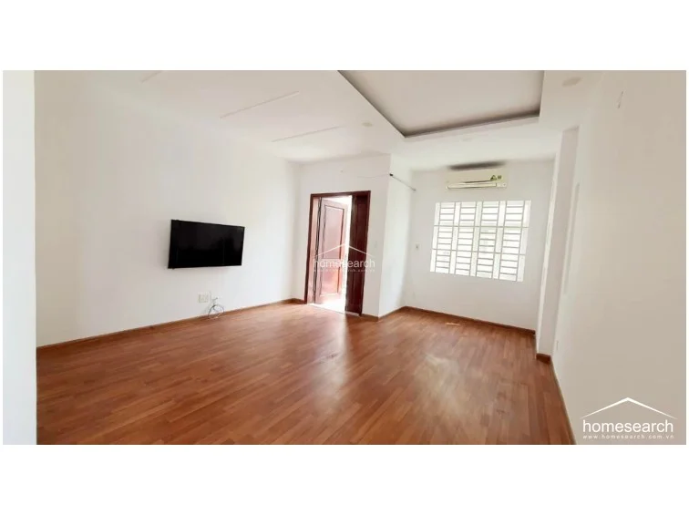 This bedroom or secondary living space is finished with high-quality wood-look flooring and clean white walls, creating a bright and airy atmosphere. It includes a wall-mounted television, a split-system AC unit, and direct access to an outdoor balcony through a sturdy wooden door.