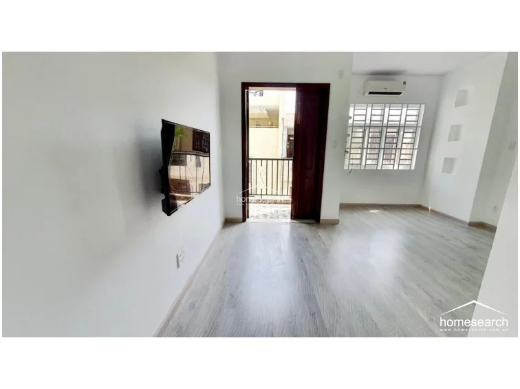 This room showcases a sleek, modern aesthetic with light-toned wood-look flooring and a minimalist wall-mounted television setup. The space is well-ventilated with a wall-mounted AC unit and features a recessed wall niche for display, leading directly out to a private balcony.