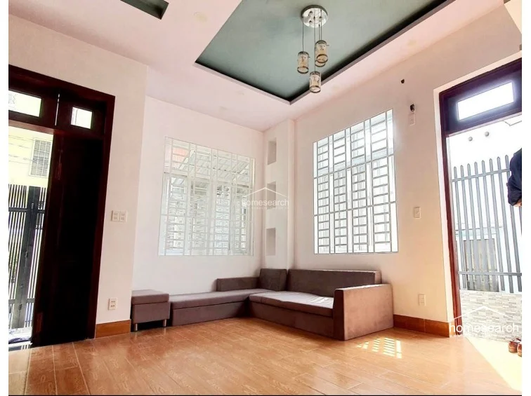 This living room maintains a consistent design language with the previous images, utilizing wood-look tiles and a recessed ceiling feature with a contemporary three-bulb chandelier. The room feels secure and well-maintained, featuring large windows with white-framed security bars that allow for ample natural light.