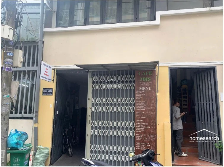 The exterior features a multi-use facade with a retractable metal security gate, currently serving as a small commercial storefront with a menu board for a cafe. The structure appears to be a modest, multi-story urban dwelling with a neutral beige paint finish and basic utility connections visible on the street side.