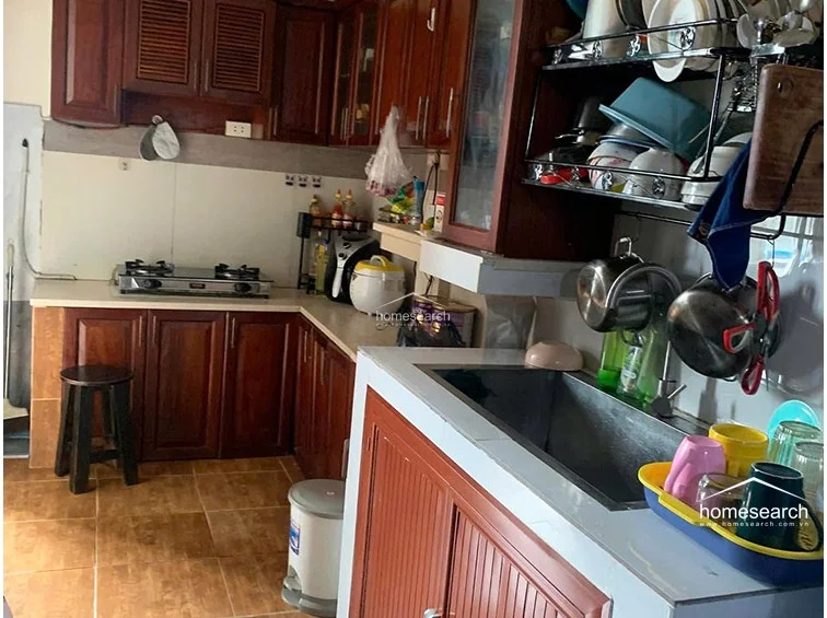 The kitchen is equipped with warm-toned wooden cabinetry and a light-colored countertop, offering a compact yet practical layout for food preparation. It includes a built-in gas stovetop and open shelving for dish storage, though the space is currently cluttered with various kitchen appliances and hanging cookware.