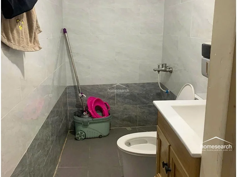 The bathroom is finished in a clean, two-tone grey tile scheme, featuring a standard white toilet and a vanity sink with a wooden base. The space is functional and well-maintained, though it currently serves as a storage area for cleaning supplies and household items.