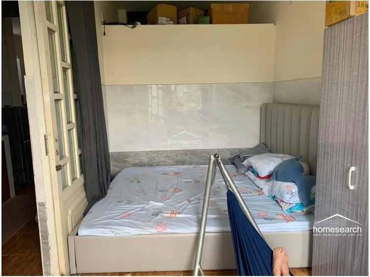 This bedroom features a platform bed with a soft grey upholstered headboard and a neutral color palette, creating a cozy sleeping environment. A tall, grey wardrobe provides essential storage, while the room connects directly to other living areas through a classic wooden-framed glass door.