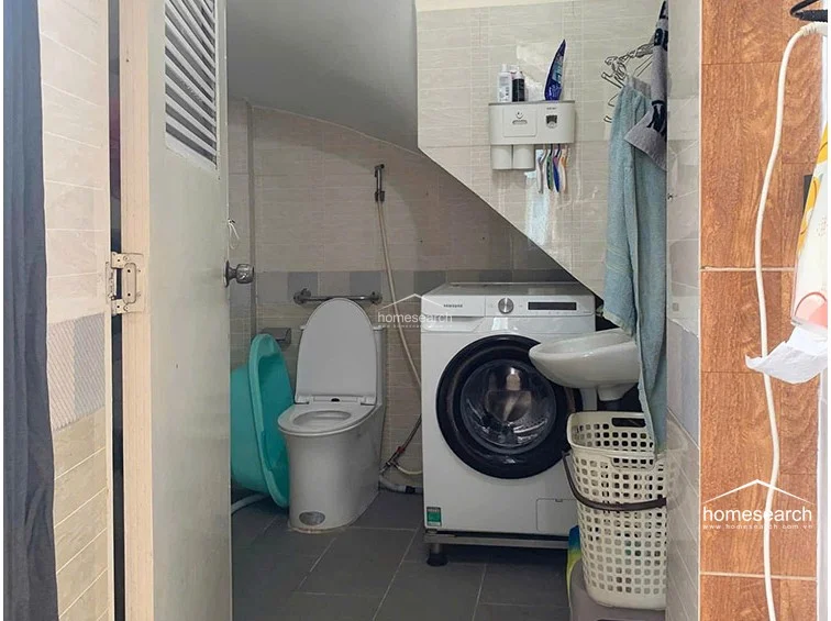 This compact utility bathroom features a functional layout that integrates a Samsung front-loading washing machine beneath a sloped ceiling, maximizing the available footprint. The space is finished with neutral-toned wall tiles and dark floor tiles, and includes a standard toilet, a wall-mounted sink, and a handheld bidet sprayer. While the area is highly utilitarian and shows signs of daily use, it offers efficient plumbing and appliance placement for a small-scale living environment.