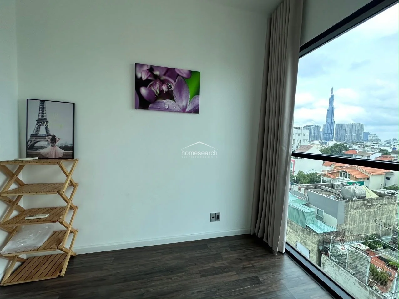 This bedroom features dark wood-look flooring and crisp white walls, creating a modern, minimalist aesthetic. A large floor-to-ceiling window with neutral curtains offers an impressive urban skyline view, while a simple wooden shelving unit provides open storage.