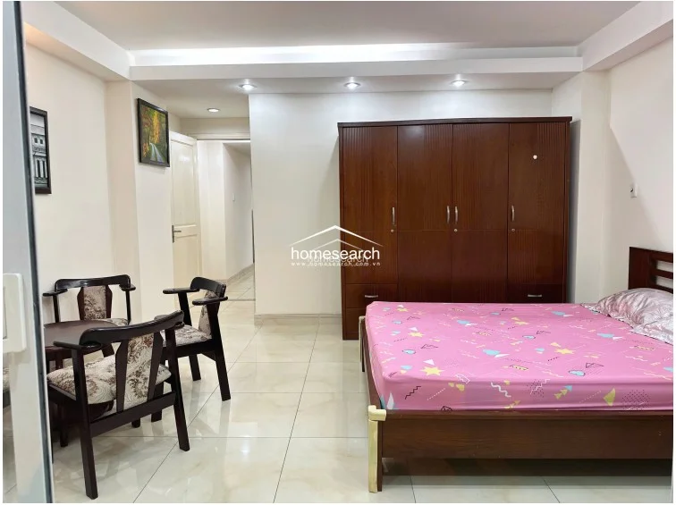 This bedroom is equipped with a large, dark-wood wardrobe providing ample storage and a bed with a vibrant pink patterned cover. The room maintains a simple, functional design with white walls, light tile flooring, and a small wooden seating area near the doorway.