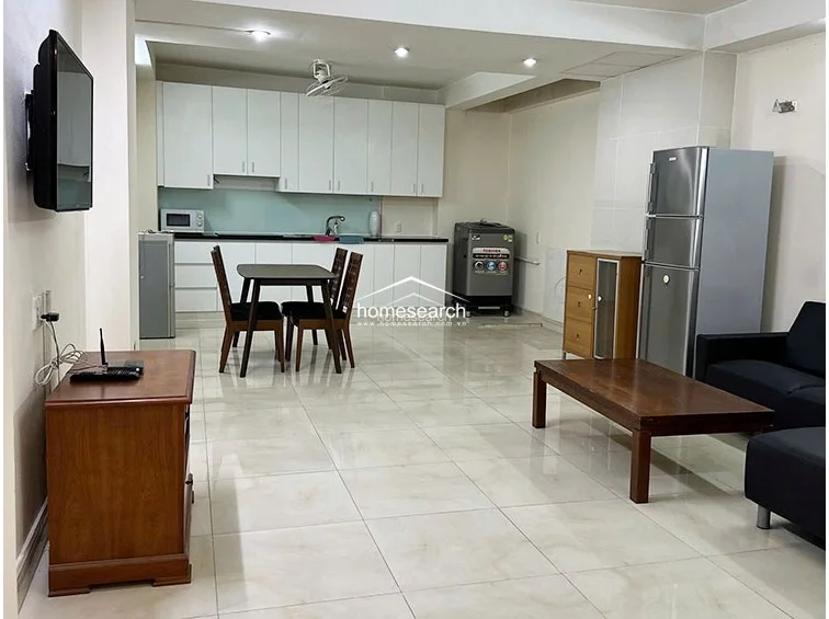 This open-concept living and kitchen area features large, light-colored floor tiles and a neutral white color scheme that enhances the sense of space. The kitchen is equipped with white cabinetry, a dark countertop, and a microwave, while the living area includes a wooden TV console and a black sofa, all maintained in good condition.