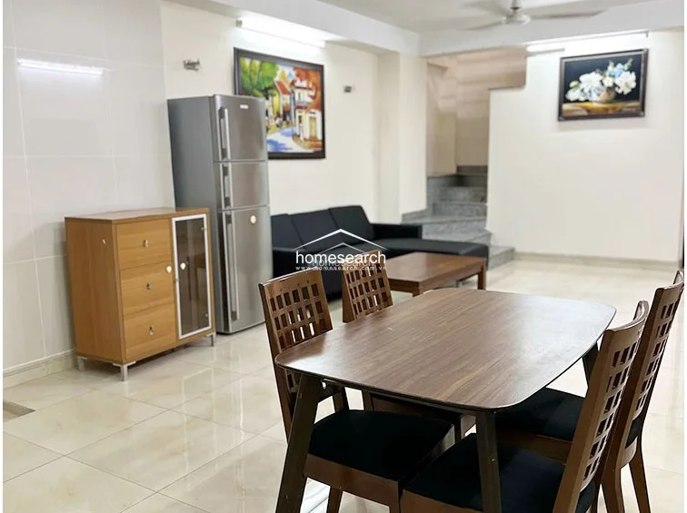 This perspective highlights the dining area, which connects directly to the living space and a staircase leading to other levels. The room is decorated with framed wall art and features a wooden dining set, maintaining a consistent, clean aesthetic throughout the open floor plan.