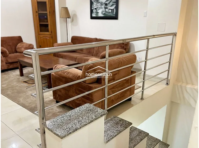 This shot shows a mezzanine or upper-level landing area overlooking a lounge space furnished with comfortable brown sofas. The staircase is finished with granite treads and a modern stainless steel railing, highlighting a clean, contemporary transition between the different levels of the property.